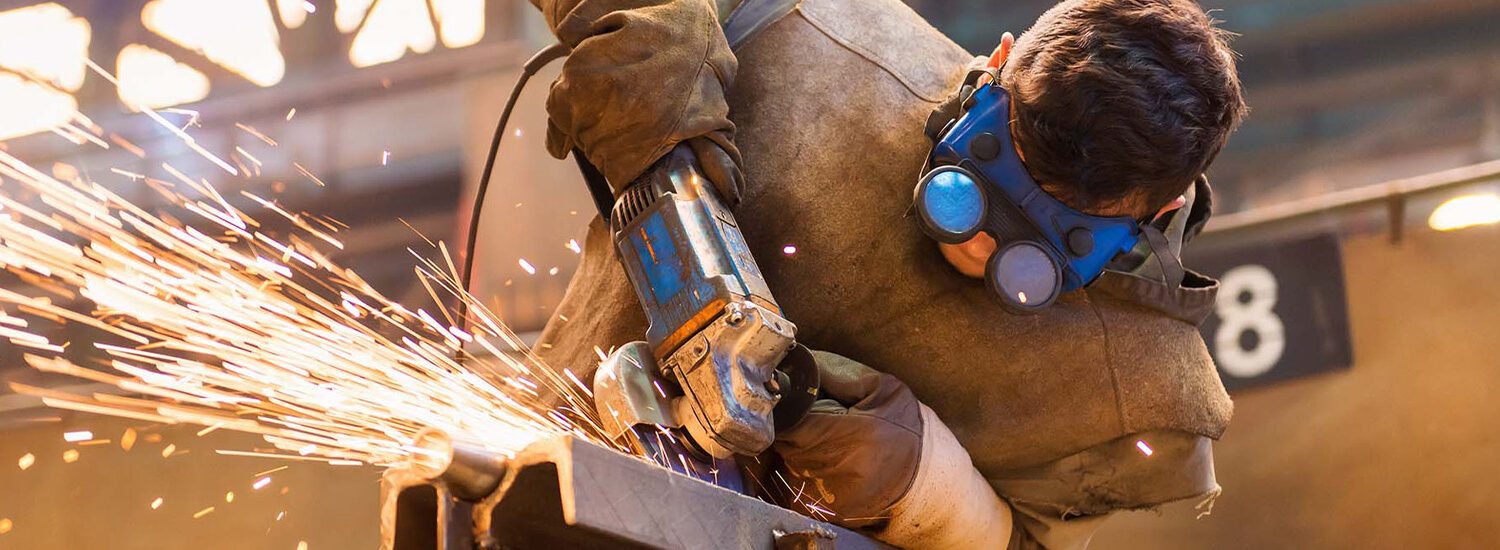 Industrial worker using a handheld grinder to cut metal with flying sparks, wearing protective gear including gloves, welding jacket, and safety goggles—illustrating heavy-duty fabrication and metalworking processes in a manufacturing facility.