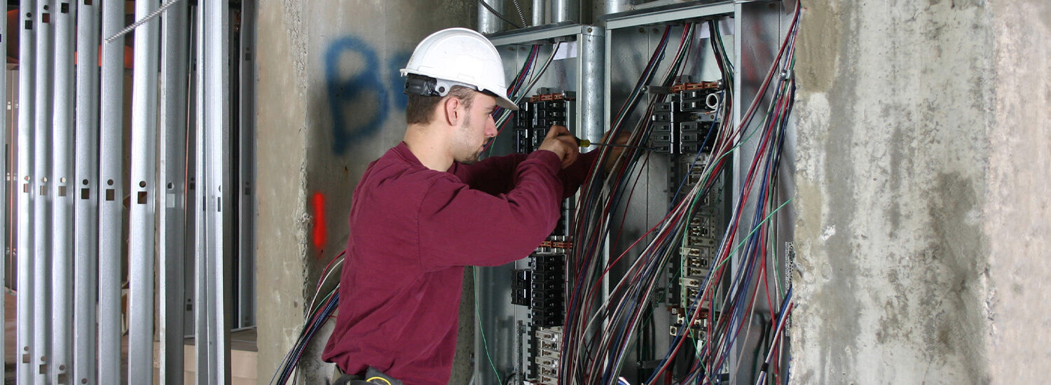 Electrician in a hard hat working on commercial electrical panel installation with exposed wiring inside an unfinished building, showcasing expert industrial electrical services and wiring solutions.