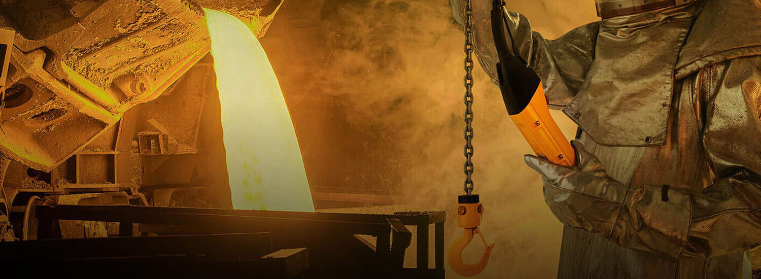Foundry worker operating a hoist near a stream of molten metal being poured, showcasing industrial lifting equipment, safety gear, and high-heat metalworking processes.