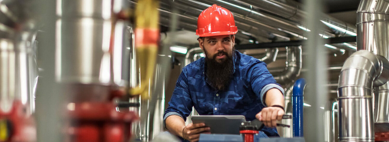 Industrial millwright technician wearing a red hard hat and safety gear operating machinery and monitoring equipment in a facility with stainless steel piping systems.