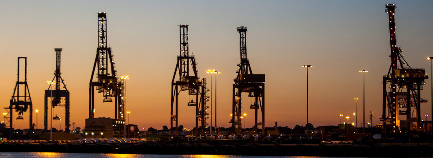 Cargo cranes silhouetted against a colorful dusk sky at a busy shipping port, highlighting global logistics, freight transportation, and industrial shipping infrastructure.